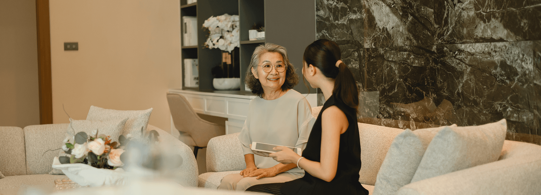 Two women having a conversation on a cream sofa in an elegant living room with marble wall and floral accents.