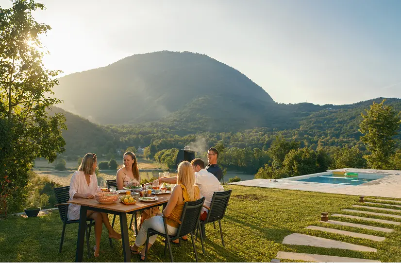 Family dining outdoors on a sunlit hillside with mountain views and pool.