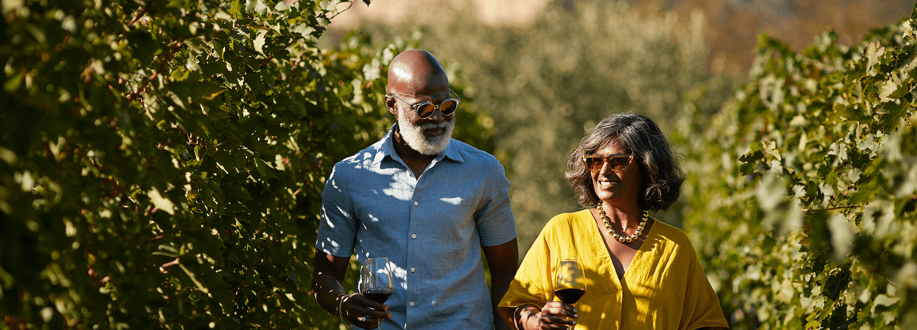 Smiling couple walking through a sunlit vineyard, holding wine glasses and enjoying the outdoors together.