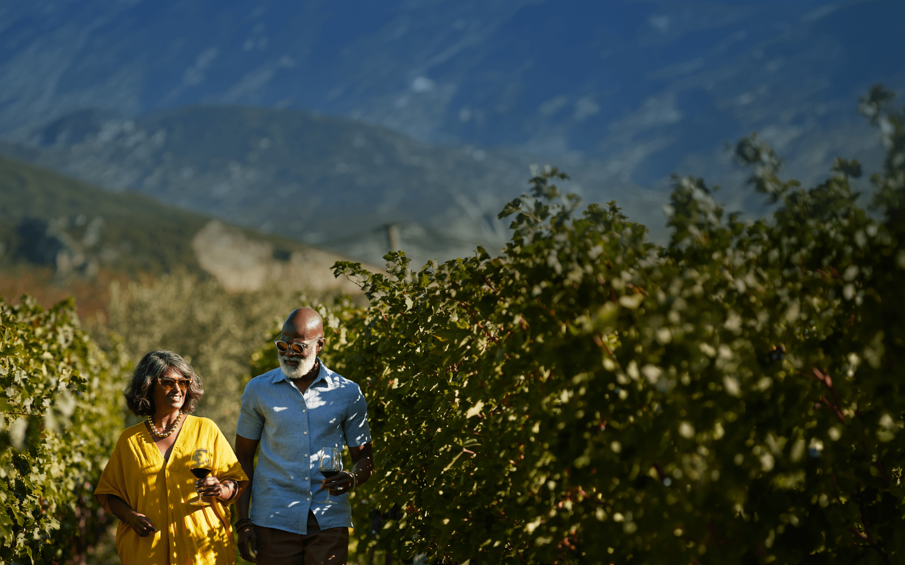 Smiling couple walking through a sunlit vineyard, holding wine glasses and enjoying the outdoors together.
