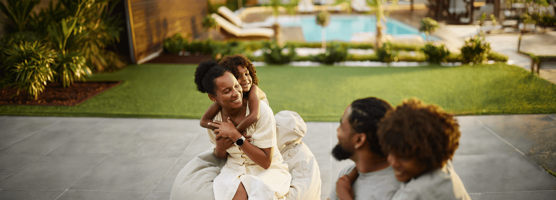 Family relaxing outdoors at golden hour — mother holding a child with another child nearby, a lush lawn and pool in the background.