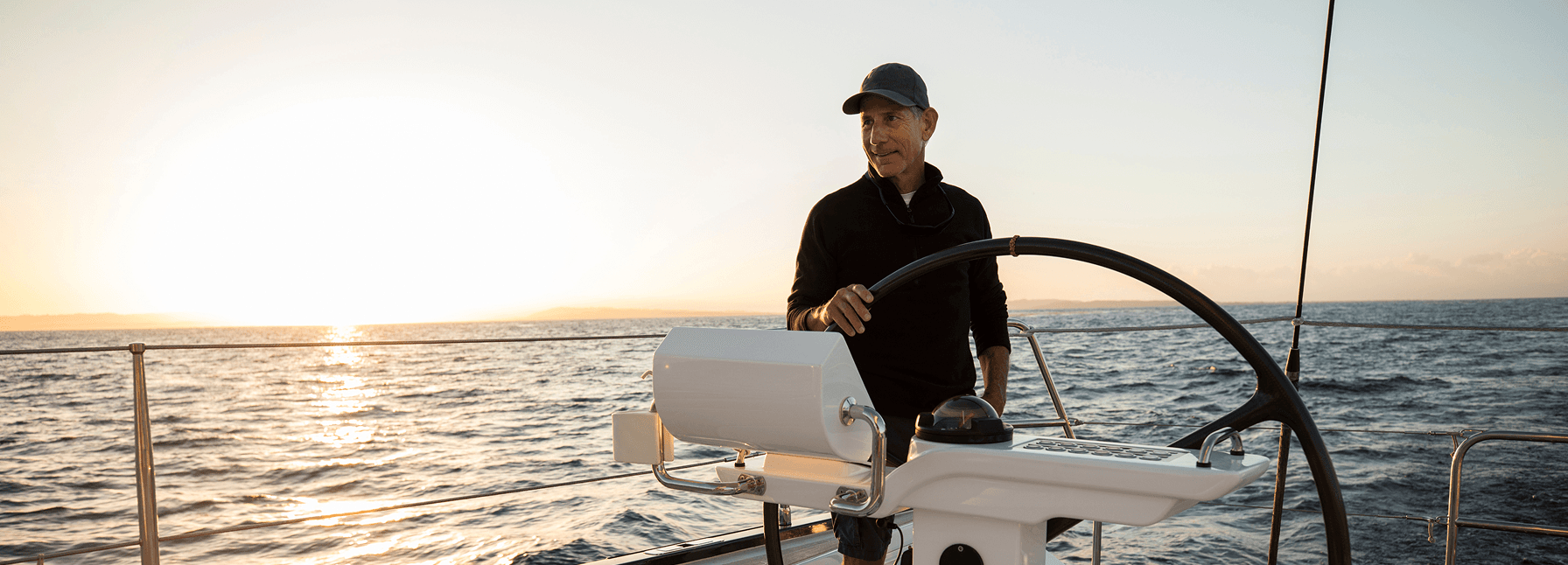 Man in a cap steering a sailboat at sunset on calm open waters.