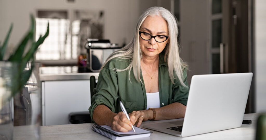 woman wearing glasses writing in notebook in front of laptop