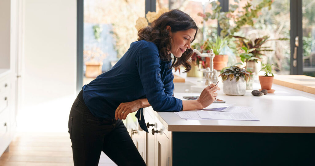 Woman standing over kitchen counter, reviewing documents.