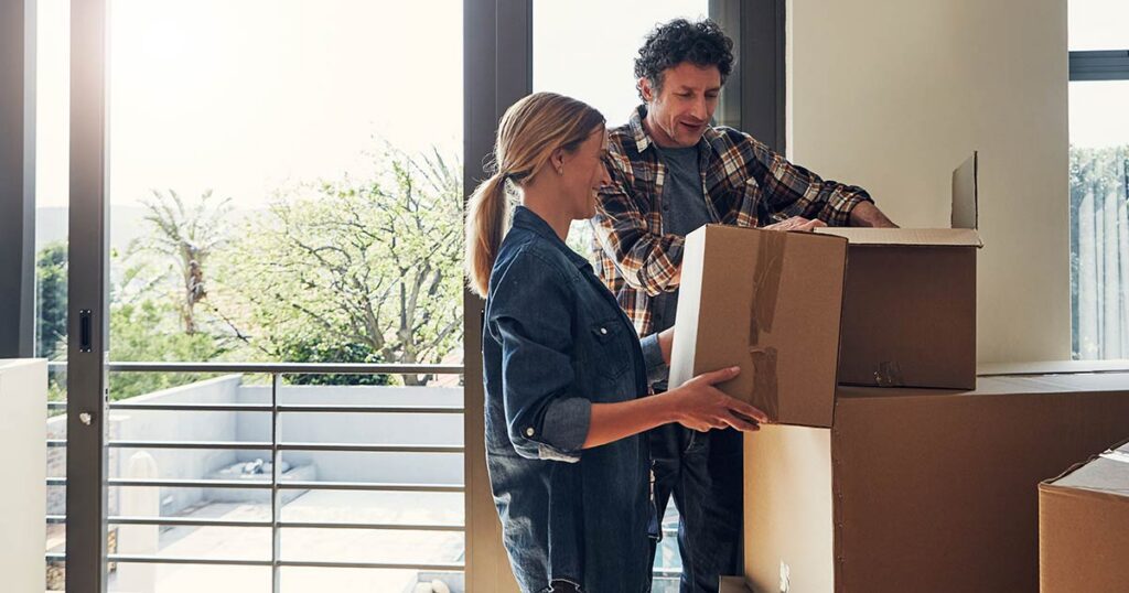 Couple unpacking cardboard moving boxes in a new home overlooking a balcony.