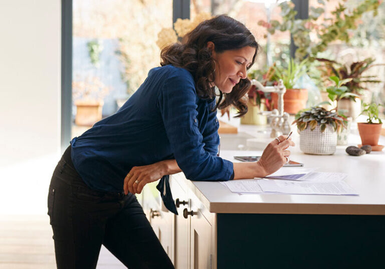 Woman standing over kitchen counter, reviewing documents.