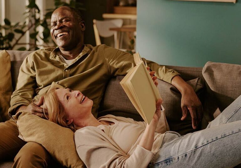 Senior couple laughing and reading on a living room sofa, representing an expat and foreign spouse planning Social Security retirement benefits.