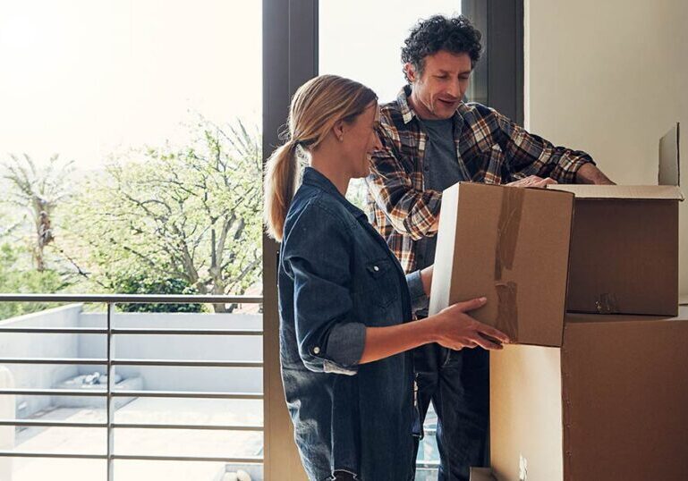 Couple unpacking cardboard moving boxes in a new home overlooking a balcony.