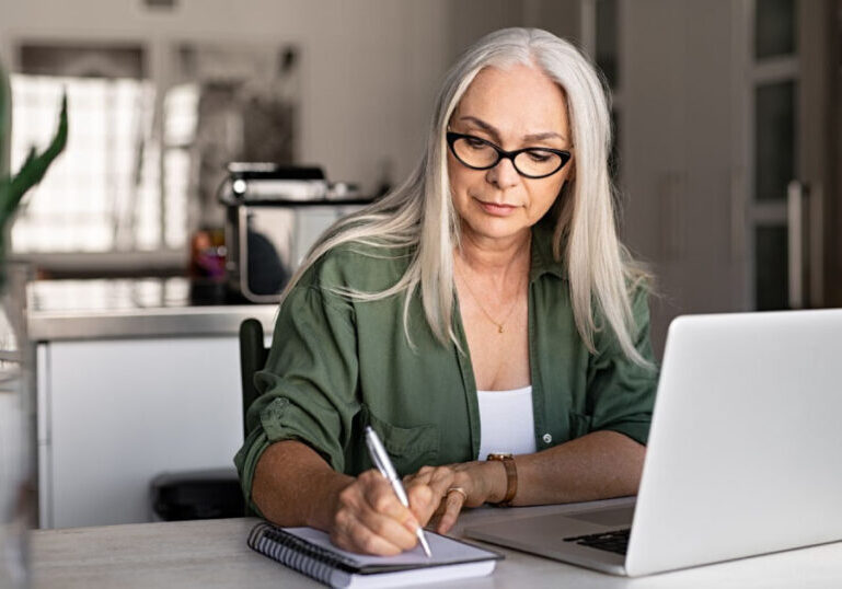 woman wearing glasses writing in notebook in front of laptop