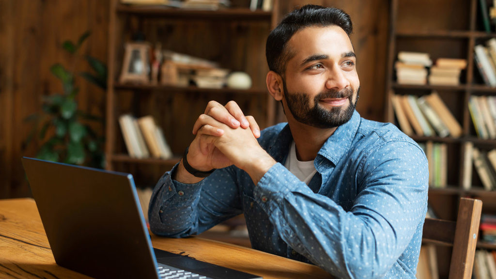 Happy and optimistic Indian man in casual wear using laptop in modern office, successful inspired arabic male entrepreneur looks away and smiles thoughtfully, planning next step