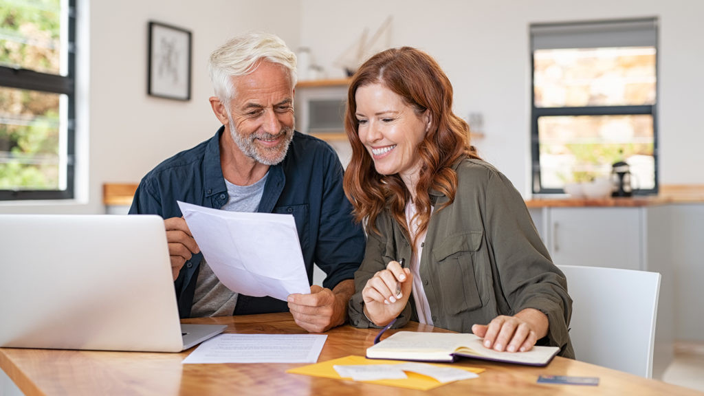 Mature smiling couple sitting and managing expenses at home. Happy senior man and mid woman paying bills and managing budget. Middle aged couple checking accountancy and bills while looking at receipt.