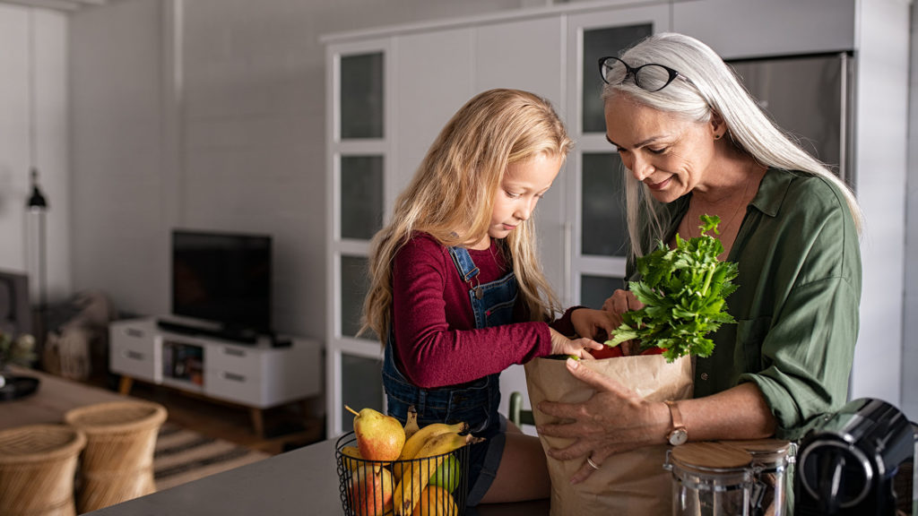 Grandmother and her granddaughter holding grocery shopping bag with vegetables at home. Happy old grandma holding paper bag with vegetables while grandchild removing food from bag. Senior lovely woman and little girl sitting on kitchen counter with groceries.
