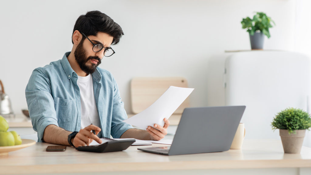 Paying bills, taxes at home online. Young arab man using calculator and laptop computer, calculating taxes or planning budget while sitting at kitchen table at home, copy space