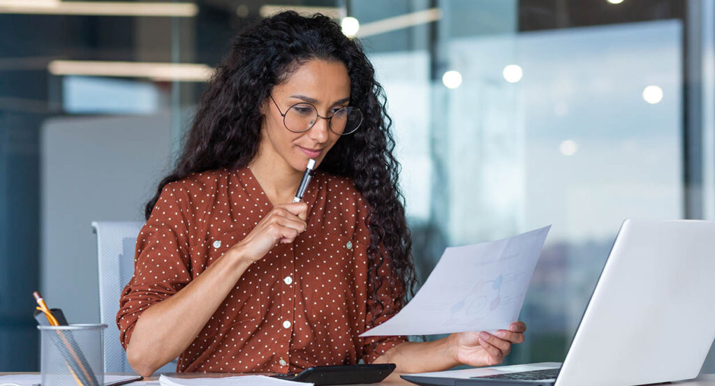 Beautiful hispanic woman in office doing paperwork, successful businesswoman financier working inside office sitting at table using laptop, at work with contracts and bills, thinking about decision.