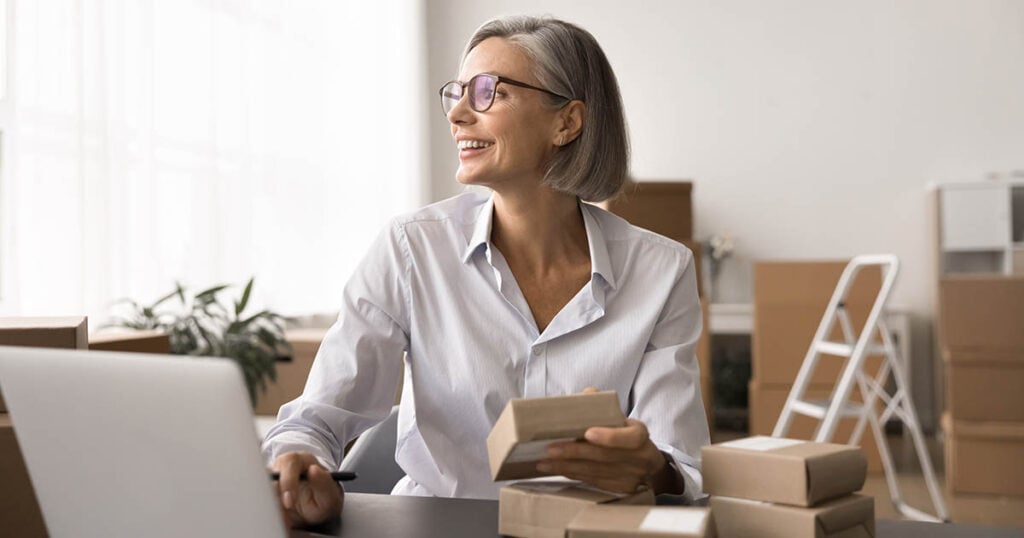 Small business owner sitting at a desk with a laptop, holding product boxes in a workspace filled with packages.