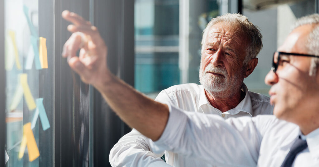 Two older professionals reviewing plans on a glass wall, representing how today’s millionaires take a more active, strategic approach to designing their retirement lifestyle