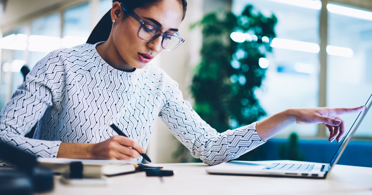 Woman taking notes while working on a laptop in an office.