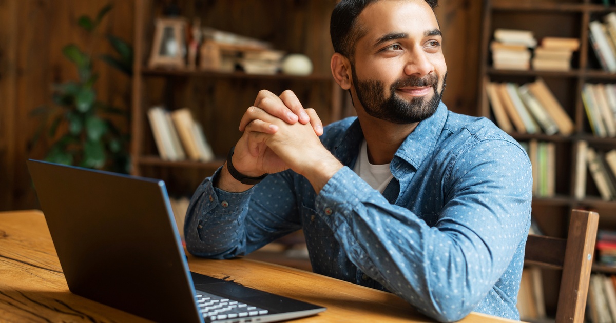 Man sitting at a desk with a laptop, smiling in a warm office setting.