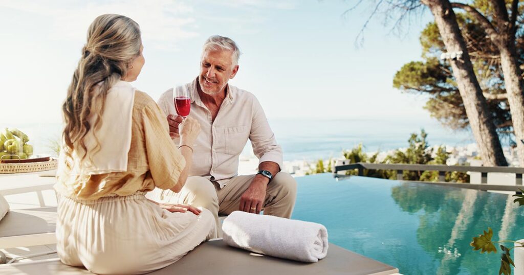 A retired senior couple smiling and toasting drinks by a luxury poolside overlooking the ocean during early retirement.