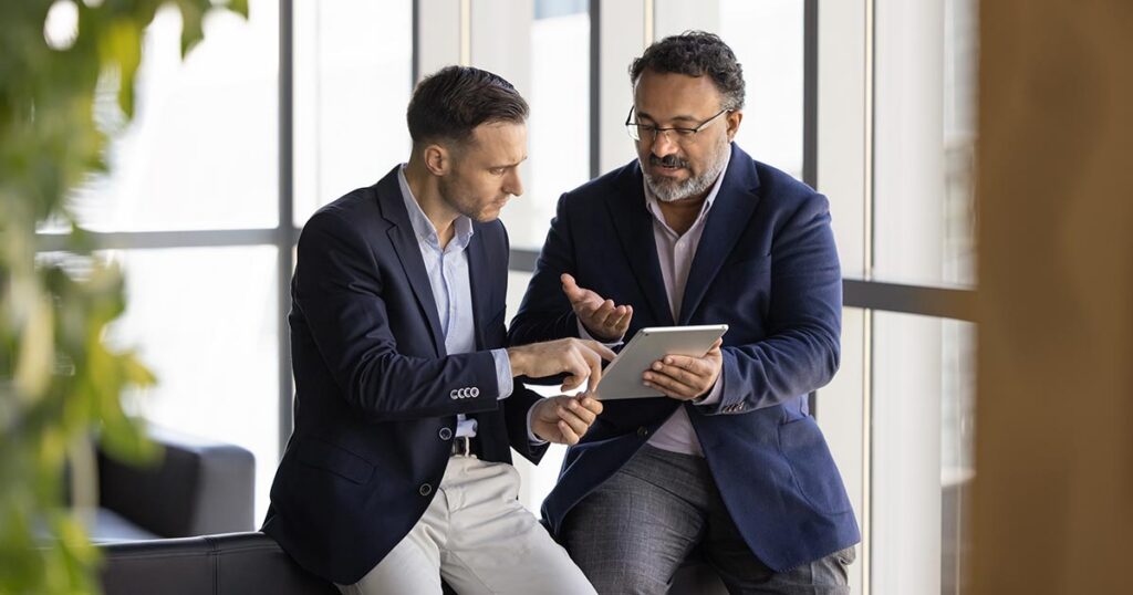 Two business finance professionals in suits reviewing balance sheet data on a digital tablet in an office.