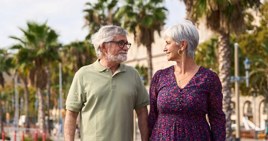 Retired couple smiling and walking outdoors along a palm tree-lined sidewalk, illustrating considerations for retirement relocation.