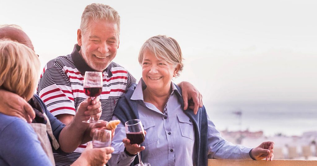 A smiling group of older adult friends socializing and toasting with glasses of red wine outdoors.