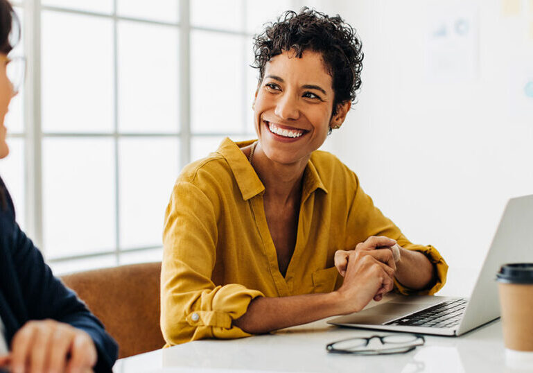 Two colleagues smiling and discussing retirement planning over a laptop at an office desk.