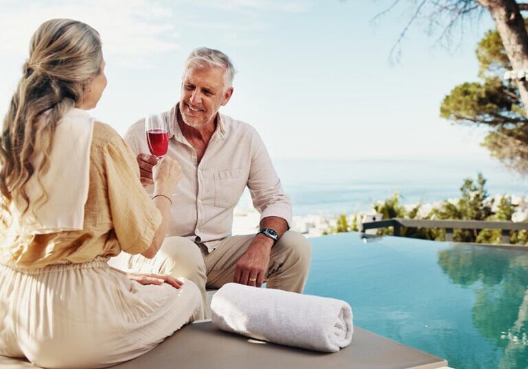 A retired senior couple smiling and toasting drinks by a luxury poolside overlooking the ocean during early retirement.