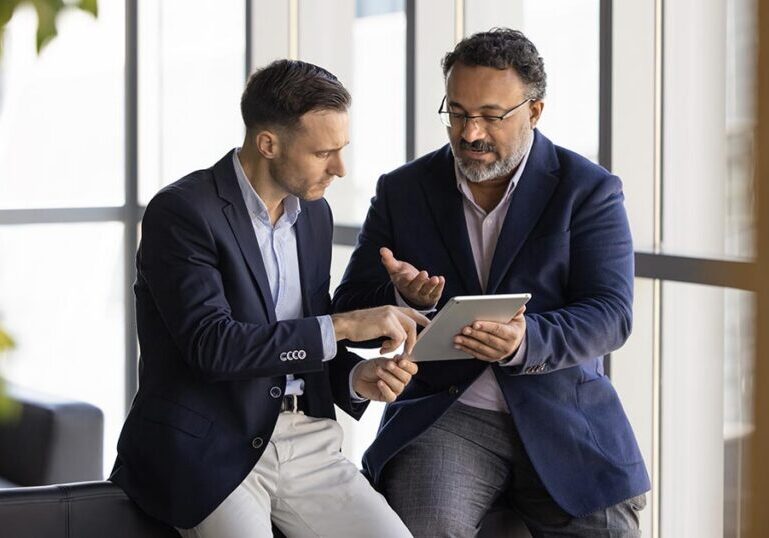 Two business finance professionals in suits reviewing balance sheet data on a digital tablet in an office.