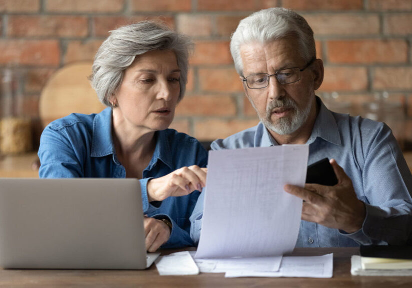 Mature couple compares notes between paperwork and laptop