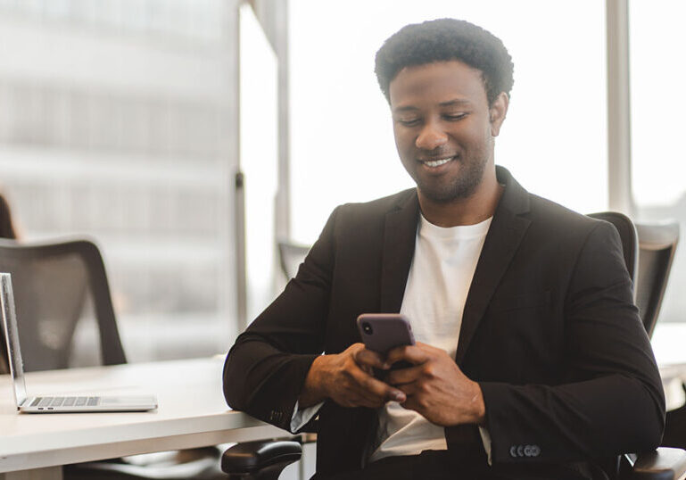 Attractive middle aged african american man holding mobile phone reading text message in modern office. Pensive guy sitting with smartphone using mobile app shopping online, ordering food