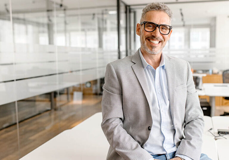 Successful good-looking modern mature businessman leaned on the desk in contemporary office, middle-aged male employee, small business owner, investor in suit looking at the camera, proud entrepreneur