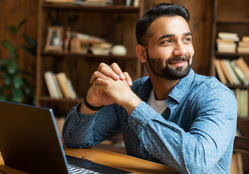 Happy and optimistic Indian man in casual wear using laptop in modern office, successful inspired arabic male entrepreneur looks away and smiles thoughtfully, planning next step