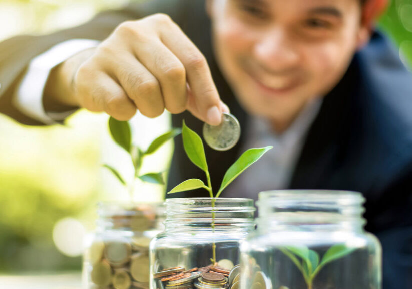 Businessman putting coin into the glass jar with young plant, demonstrating financial growth through saving plans and investment schemes