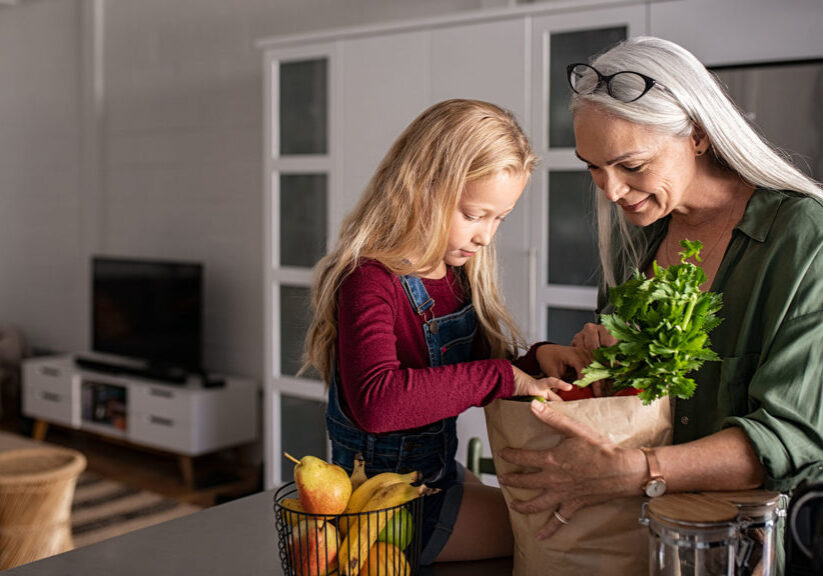 Grandmother and her granddaughter holding grocery shopping bag with vegetables at home. Happy old grandma holding paper bag with vegetables while grandchild removing food from bag. Senior lovely woman and little girl sitting on kitchen counter with groceries.