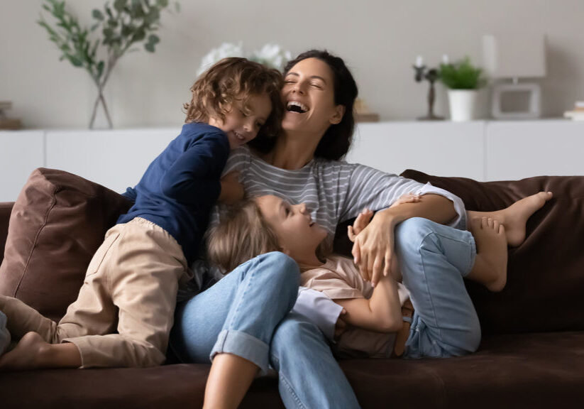 Overjoyed mother with two kids having fun on comfortable couch at home, laughing happy mom with adorable daughter and son tickling, engaged in funny active game, spending leisure time together