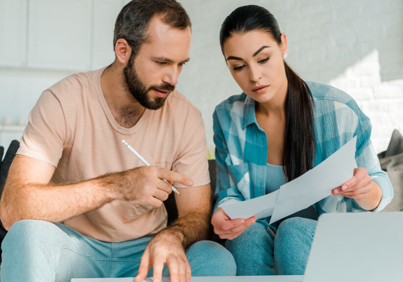 serious couple sitting on couch, filling papers, using laptop and planning