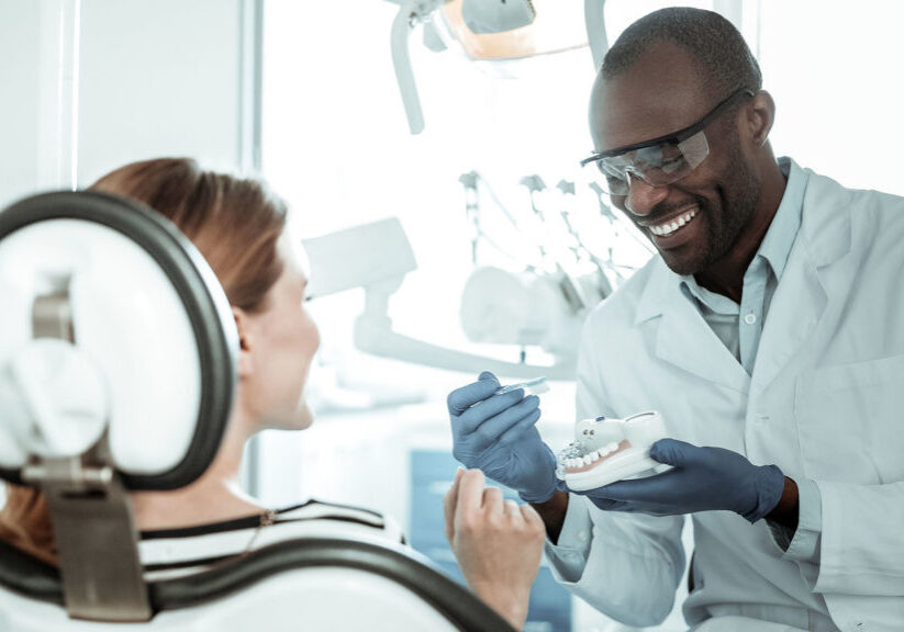 Concentrated dark-skinned doctor. Openly-smiling dentist carrying plastic model of the jaw and showing right way of brushing